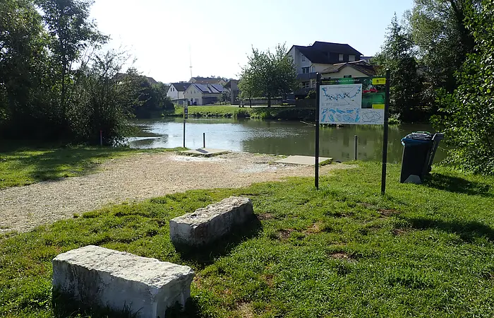 Grasfläche mit zwei Steinbänken, Infotafel und Gewässer mit Häusern im Hintergrund an sonnigem Tag