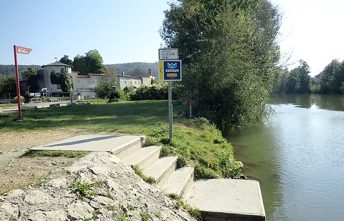 Ufer mit Treppe und Hinweisschildern an einem Fluss, im Hintergrund Gebäude und Bäume bei klarem Himmel.