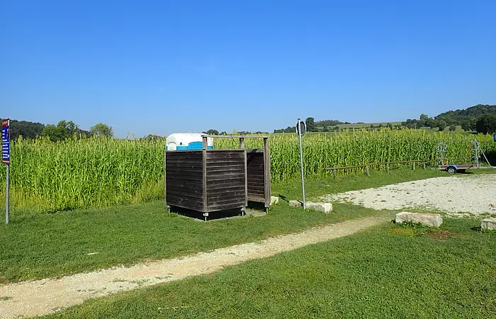 Freistehende Holzkabine mit zwei mobilen Toiletten vor einem Maisfeld unter blauem Himmel.