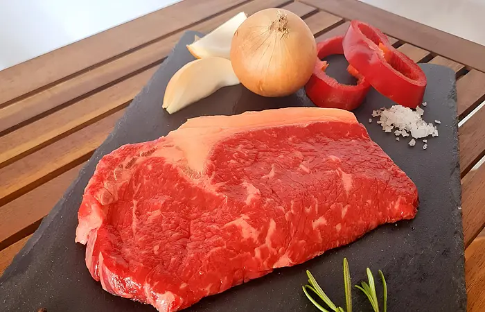 Rohe Rumpsteak mit Zwiebel, Knoblauch, Paprika, Rosmarin, Pfeffer und Salz auf schwarzer Steinplatte.