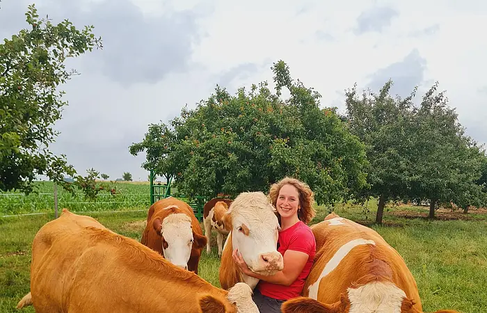 Frau in rotem Shirt umarmt Kuh auf grüner Weide mit weiteren braun-weißen Kühen und Bäumen im Hintergrund.