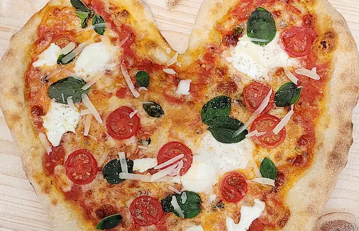 Herzförmige Pizza mit Tomatensauce, Mozzarella, Kirschtomaten, Spinat und Parmesan auf Holzuntergrund.