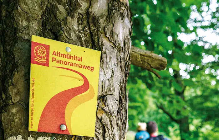 Im Vordergrund sind Wanderwegsschilder an einem Baumstamm zu sehen, daneben führt ein schmaler Pfad vorbei, auf dem zwei Wandernde unterwegs sind.