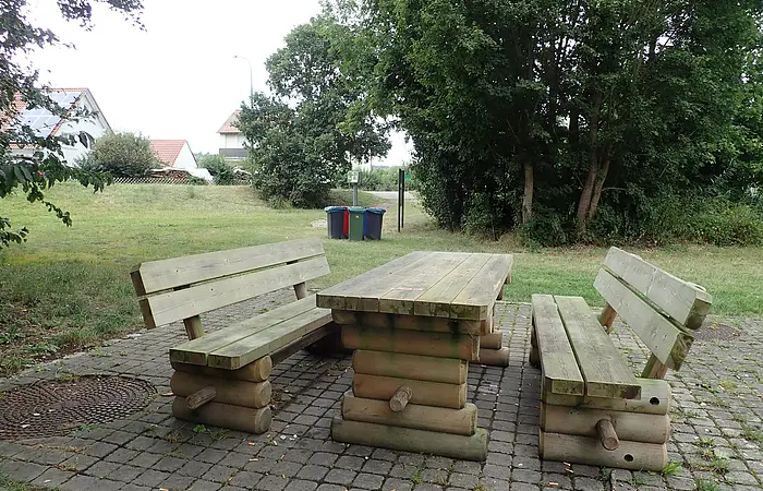 Holztisch mit zwei Holzbänken auf gepflastertem Platz im Park mit Bäumen und Häusern im Hintergrund