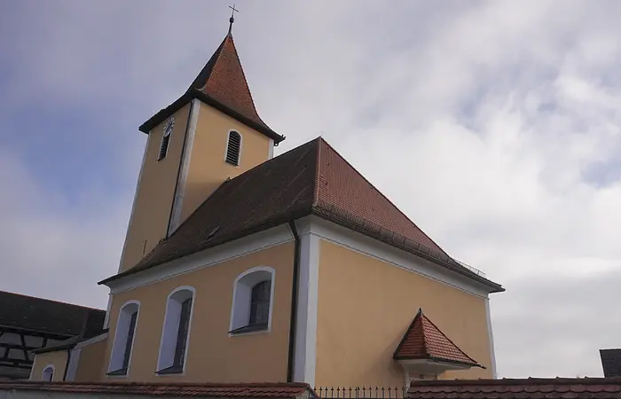 Kirchturm mit spitzem Dach und gelber Fassade unter bewölktem Himmel, von unten fotografiert.
