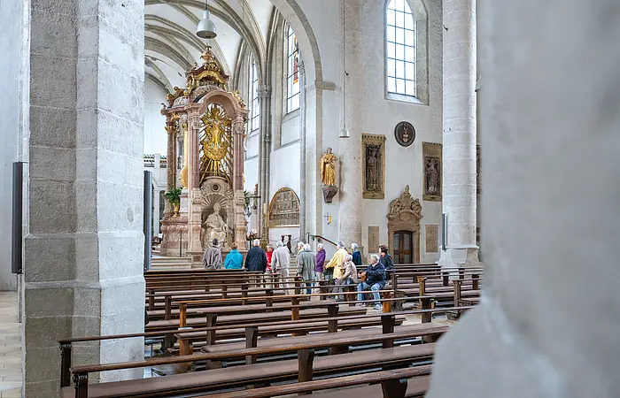 Zwischen zwei Säulen hindurch geht der Blick auf eine Gruppe, die sich in einem Gang zwischen Kirchenbänken auf einen Altar zubewegt.