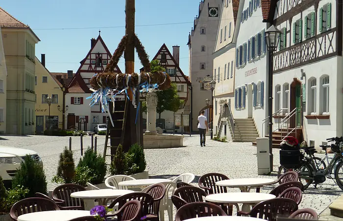 Ein Marktplatz mit Tischen und Stühlen eines Cafés im Vordergrund, Fachwerkhäusern im Hintergrund und einem Maibaum in der Mitte des Marktplatzes. Eine Person läuft auf dem Markplatz.