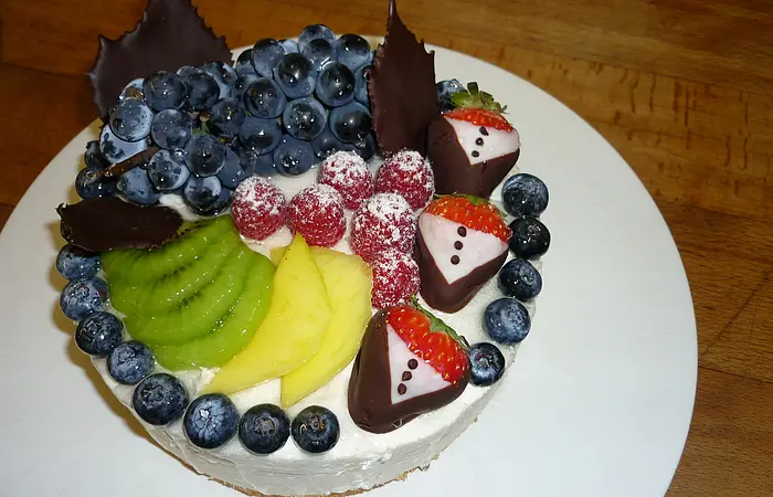 Obsttorte mit weißer Creme, obendrauf Trauben, gezuckerten Himbeeren, Kiwi, Ananas, Blaubeeren und Erdbeeren in Schokoladenmantel auf einem weißem Teller serviert.