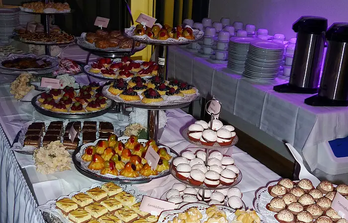 Buffet mit verschiedenen süßen Kuchen, Gebäck und Obsttörtchen auf mehreren Etageren und Tellern auf einem Tisch aufgedeckt. Im Hintergrund sind aufgestapelte beleuchtete Teller und Tassen mit zwei Kaffeekochern.