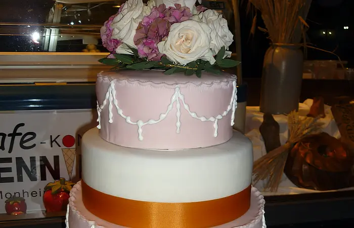 Vierstöckige Hochzeitstorte mit weißer und rosa Glasur, orangene-gelbem Band um zwei Stockwerke und einem rosa-weißen Blumenarrangement obenauf.