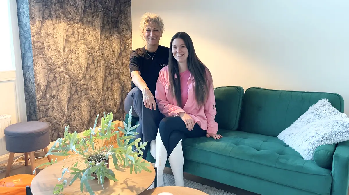 Bahar und Alex Zwei Yogalehrerinnen sitzen auf einem grünen Sofa. Im Vordergrund steht ein runder Tisch mit einer grünen Pflanze.