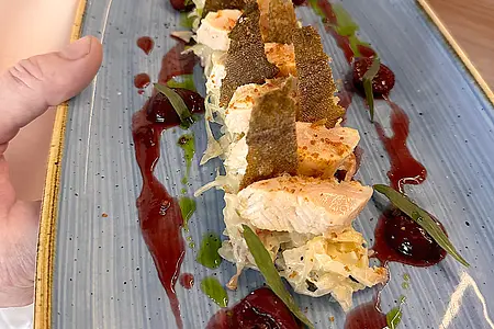 Blauer rechteckiger Teller mit angerichtetem Fischfilet, grünen Blättern und roten Soßentropfen, gehalten von zwei Händen.