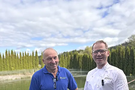Zwei Männer stehen an einem Teich, einer trägt ein blaues Poloshirt, der andere eine weiße Kochjacke.