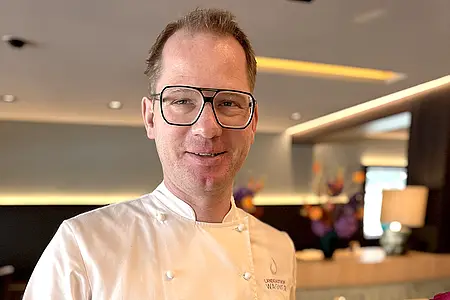 Mann in Kochjacke mit Brille hält Teller mit angerichtetem Gericht in Restaurantumgebung.