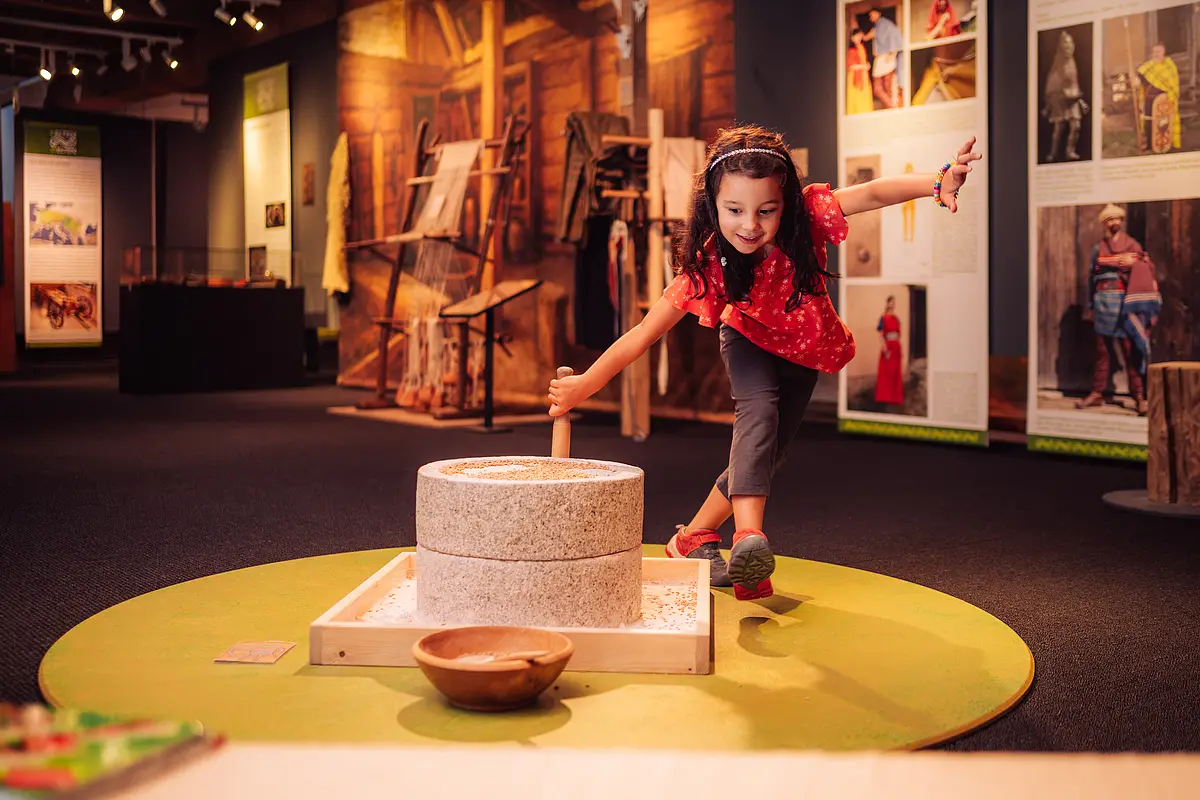 Mädchen bedient eine traditionelle Mühle in einem Museum mit Ausstellungsstücken und Informationstafeln.