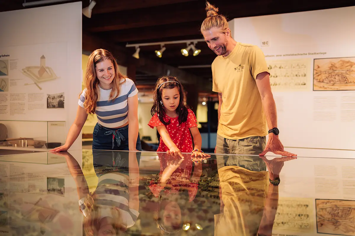 Drei Personen betrachten ein Modell hinter Glas in einem Museum; ein Mädchen zeigt auf etwas im Modell.