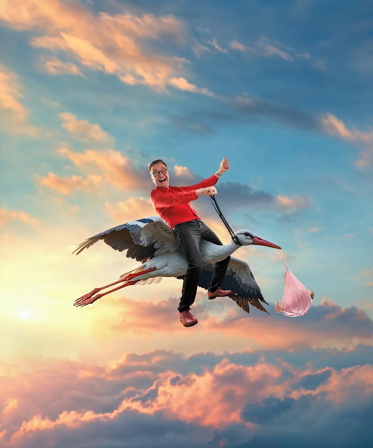 Pressebild Heinrich del Core Juhuu meine Frau wird Oma Mann in rotem Hemd reitet auf fliegendem Storch, der ein Baby in rosa Tuch trägt, vor Sonnenuntergangshimmel. Pressebild zur Veranstaltung Heinrich Del Core Juhuu meine Frau wird Oma am 09.04.2027 um 20.00 Uhr in der Stadthalle Monheim