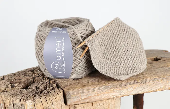 Grau-beige Merinowolle Knäuel mit Strickstück auf rustikalem Holzbrett und Holzstuhl im Hintergrund.