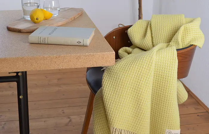 Gelbe Decke auf Holzstuhl neben Tisch mit Buch, Glas Wasser, Schneidebrett und zwei Zitronen.