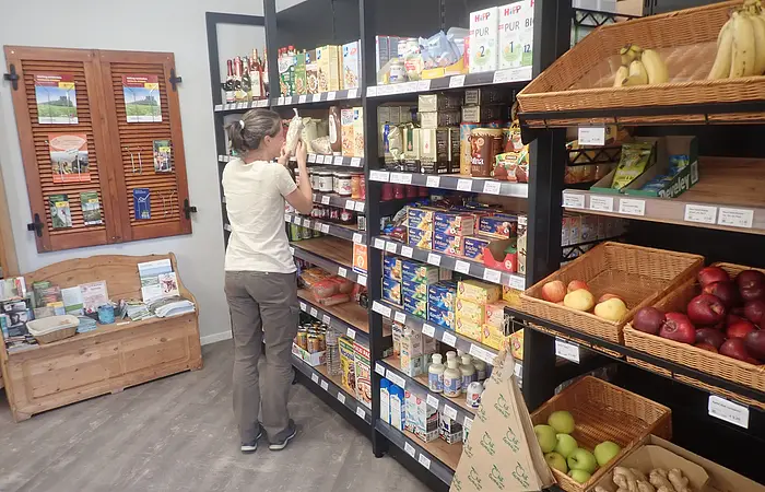 Frau steht vor einem Regal mit Lebensmitteln in einem kleinen Laden, rechts Obstkörbe mit Äpfeln und Bananen.
